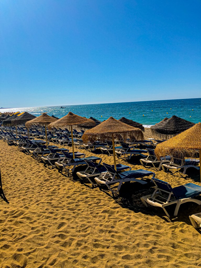 Marbella Beach in Costa del Sol Spain. Sand in front, sunbeds and umbrellas in centre and ocean in background. Blue sky.