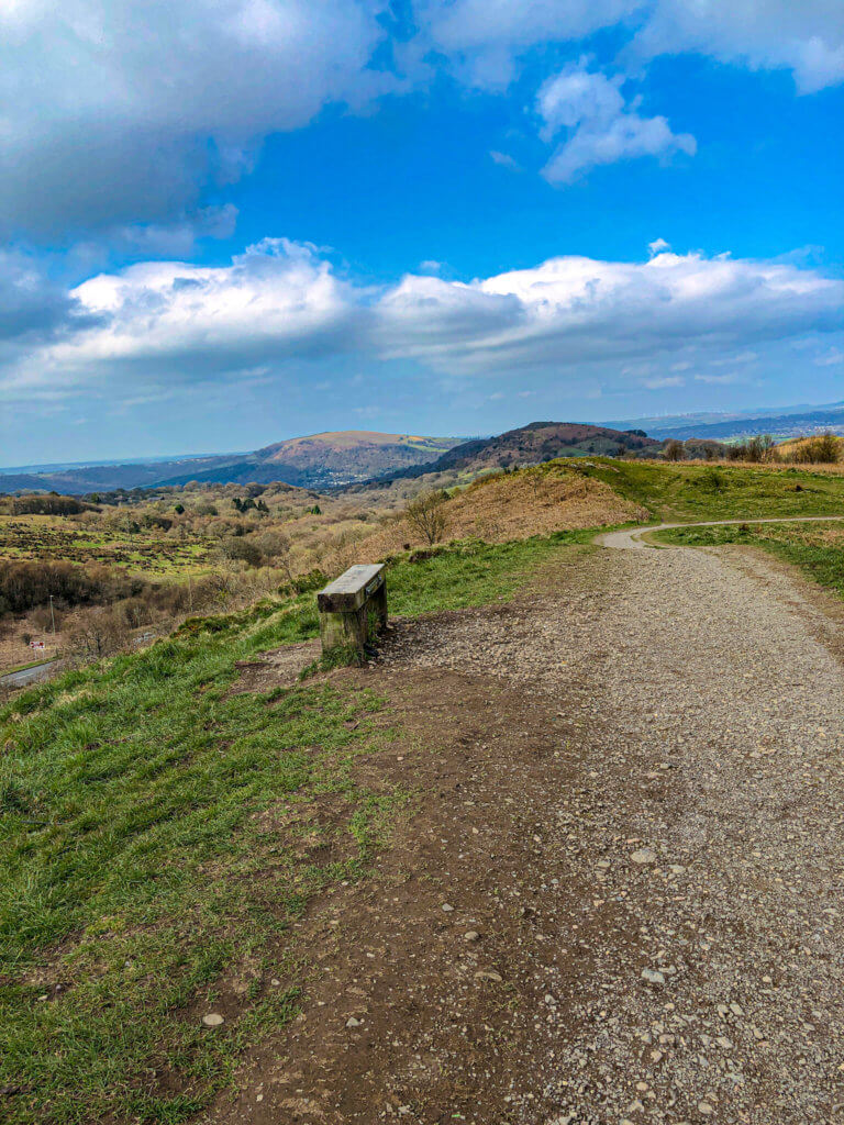 Caerphilly Mountain walks bench on left hand side with path and grass either side and blue sky in back