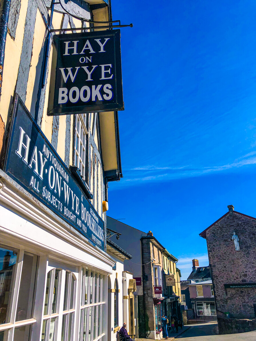 Hay on Wye Bookshops and things to do in Hay on Wye feature image of Hay on Wye black sign in left of image with blue sky and buildings on the right