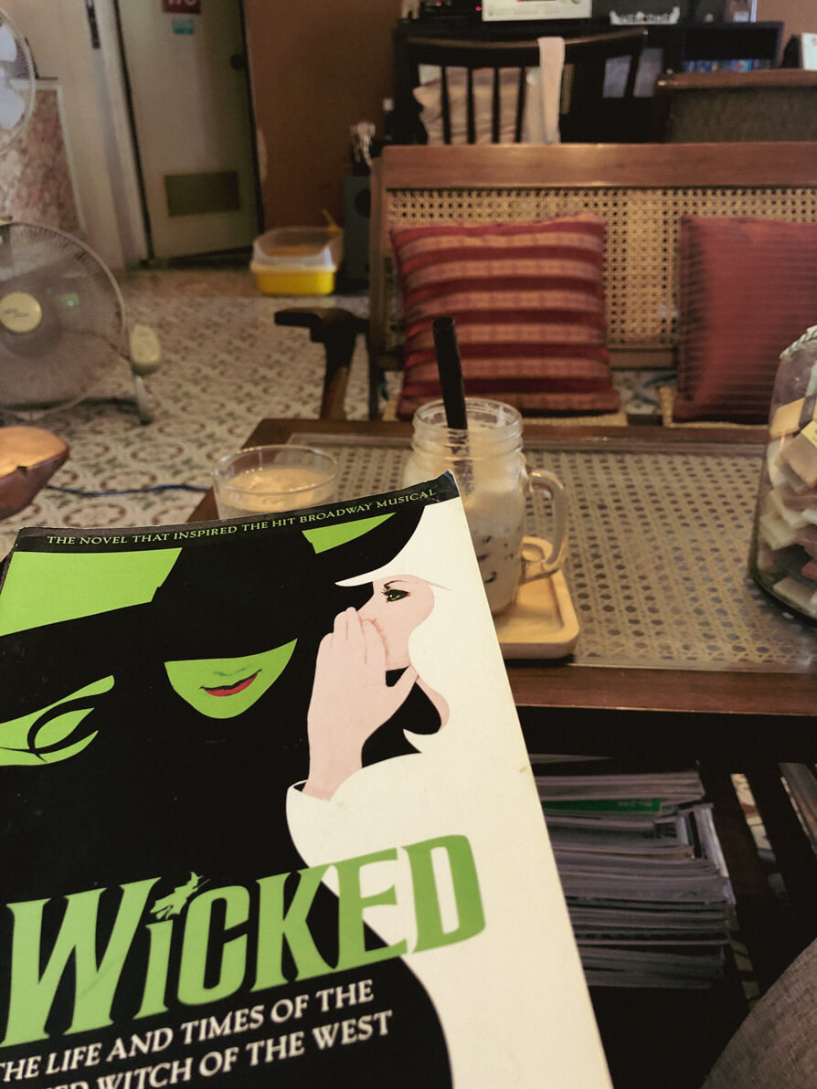 Image of Wicked book in the left side of image with cafe table, chairs and iced coffee in background
