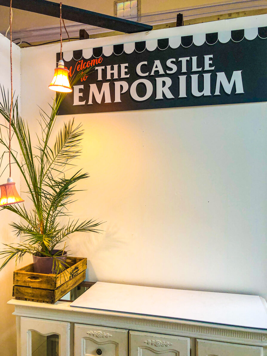 Image of The Castle Emporium sign on a white wall with white table in front. To the left there is a green, tall plant in a wooden box