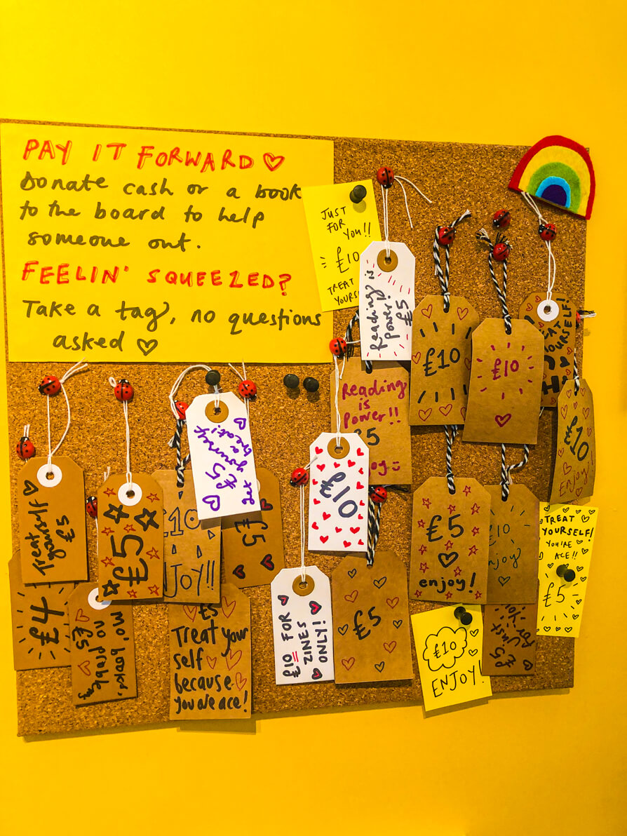 Image of Cardiff Shellife bookshop pay it forward scheme. There are several brown paper tags with gifts of '£5' or '£10' written on them. Description of the scheme reads 'Pay it Forward. Donate cash or a book for the board to help someone out. Feelin' Squeezed? Take a tag, no questions asked.'