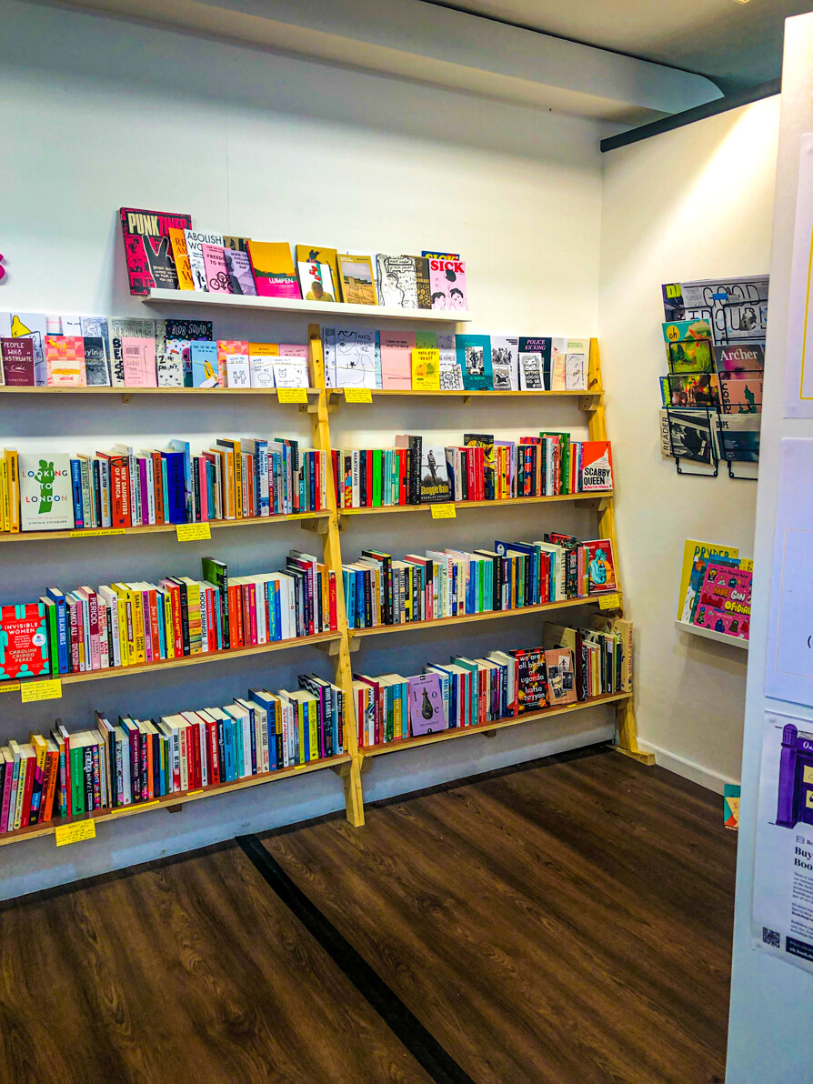 Image of interior of Cardiff Shelflife. Two wooden shelves with books stacked vertically along and a wall with magazines on hooks.