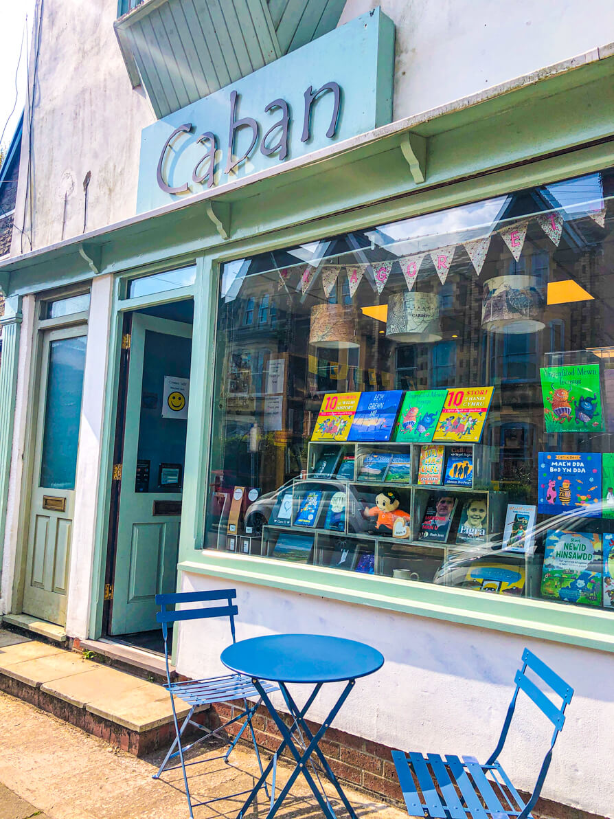 Image of the exterior of Caban bookshop in Pontcanna, Cardiff. Image shows Caban front with blue sign, blue door and window of book display. In front of the window is a set of blue chairs and table.