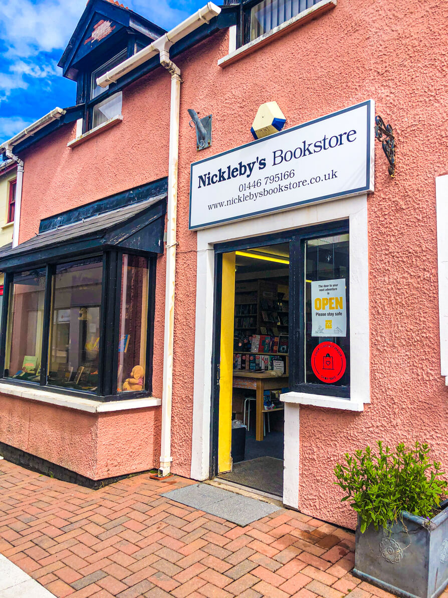 Image of exterior of Nickleby's Bookstore in Llantwit Major. The shop is painted pink with black window and display of books and door with shop sign ahead.