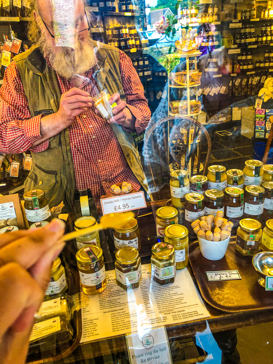 Image of Snowdonia Honey shop in Llanberis. Shop keeper with large selection of honey on a table in background and Shireen holding up stick of sample Honey in front