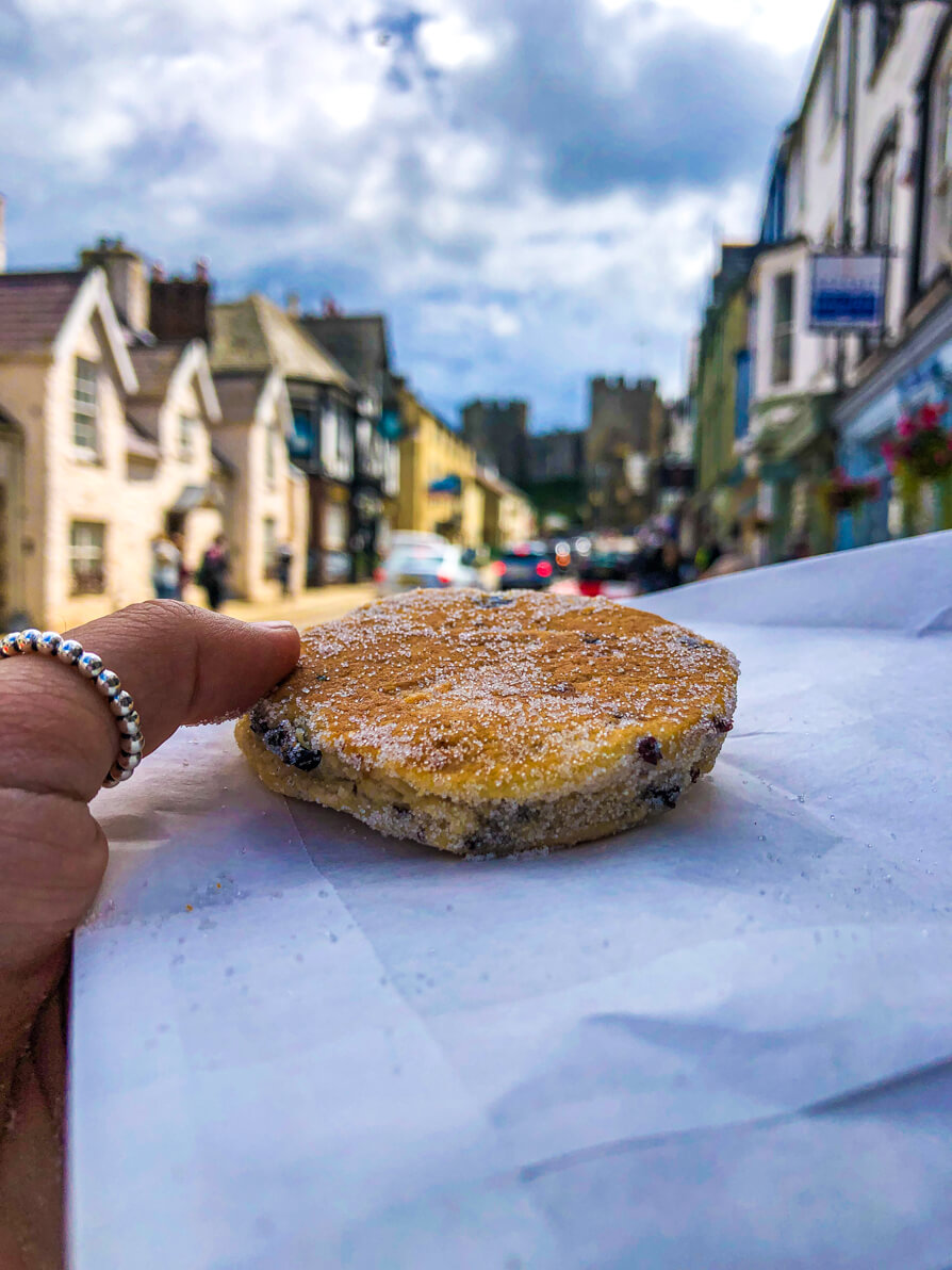 Image shows Shireen's left thumb holding up a Welsh Cake on a white paper bag with Conwy Castle in the background