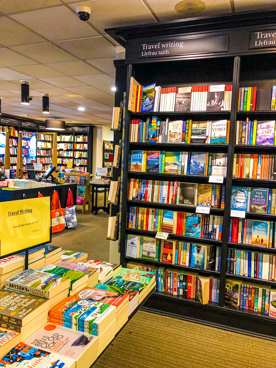 Image of Watersones Cardiff travel writing bookshelf. Black bookcase to the right of the image and table of books to the left.