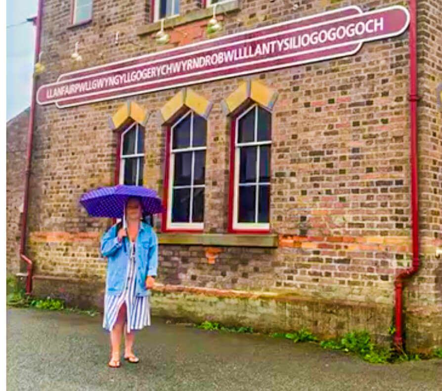 Image of Shireen with umbrella and building adjacent to train station with long Welsh word in centre on Burgundy banner