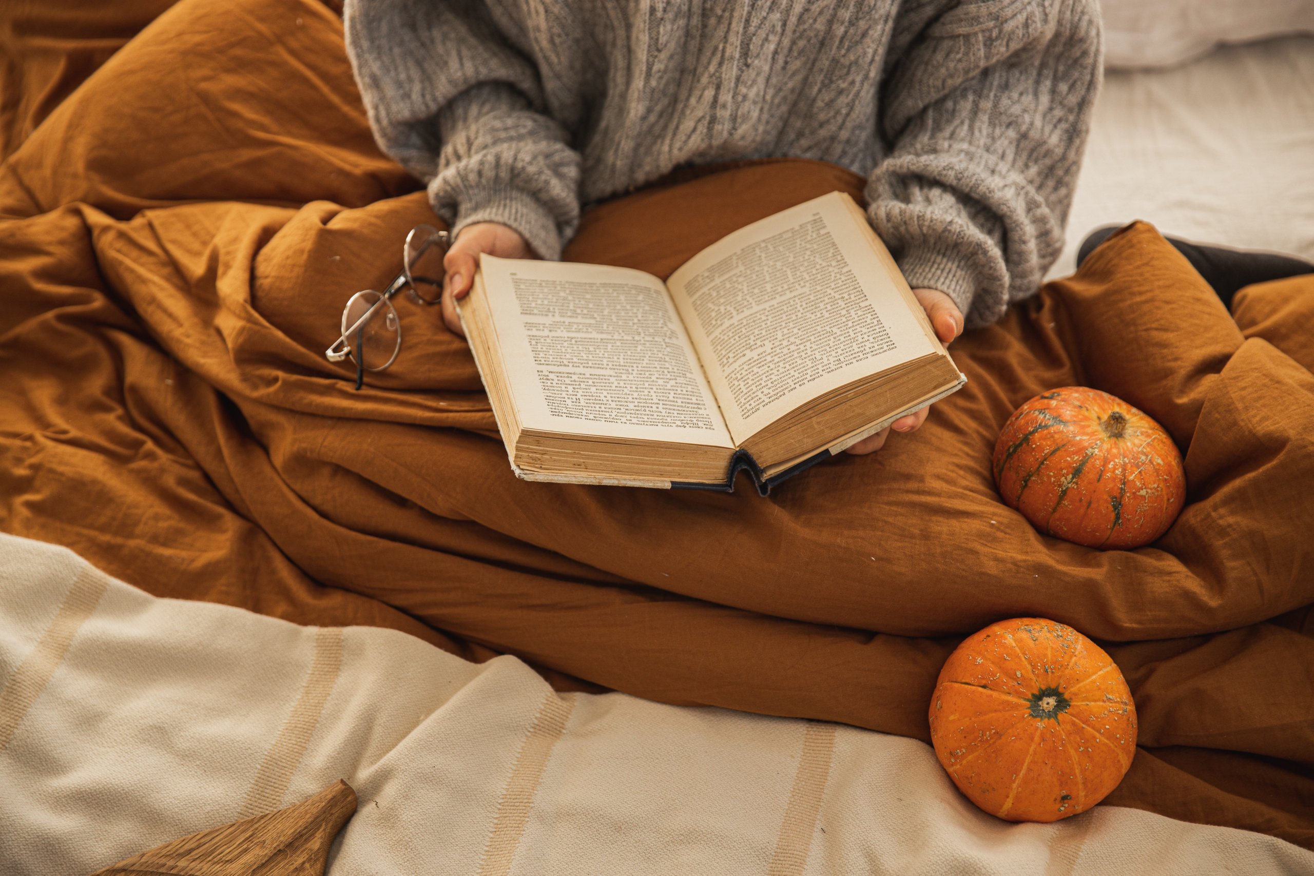 Image shows a person in grey jumper with a brown blanket sitting on a white bed reading an open book with orange pumpkins to the right. Image credit to pexels