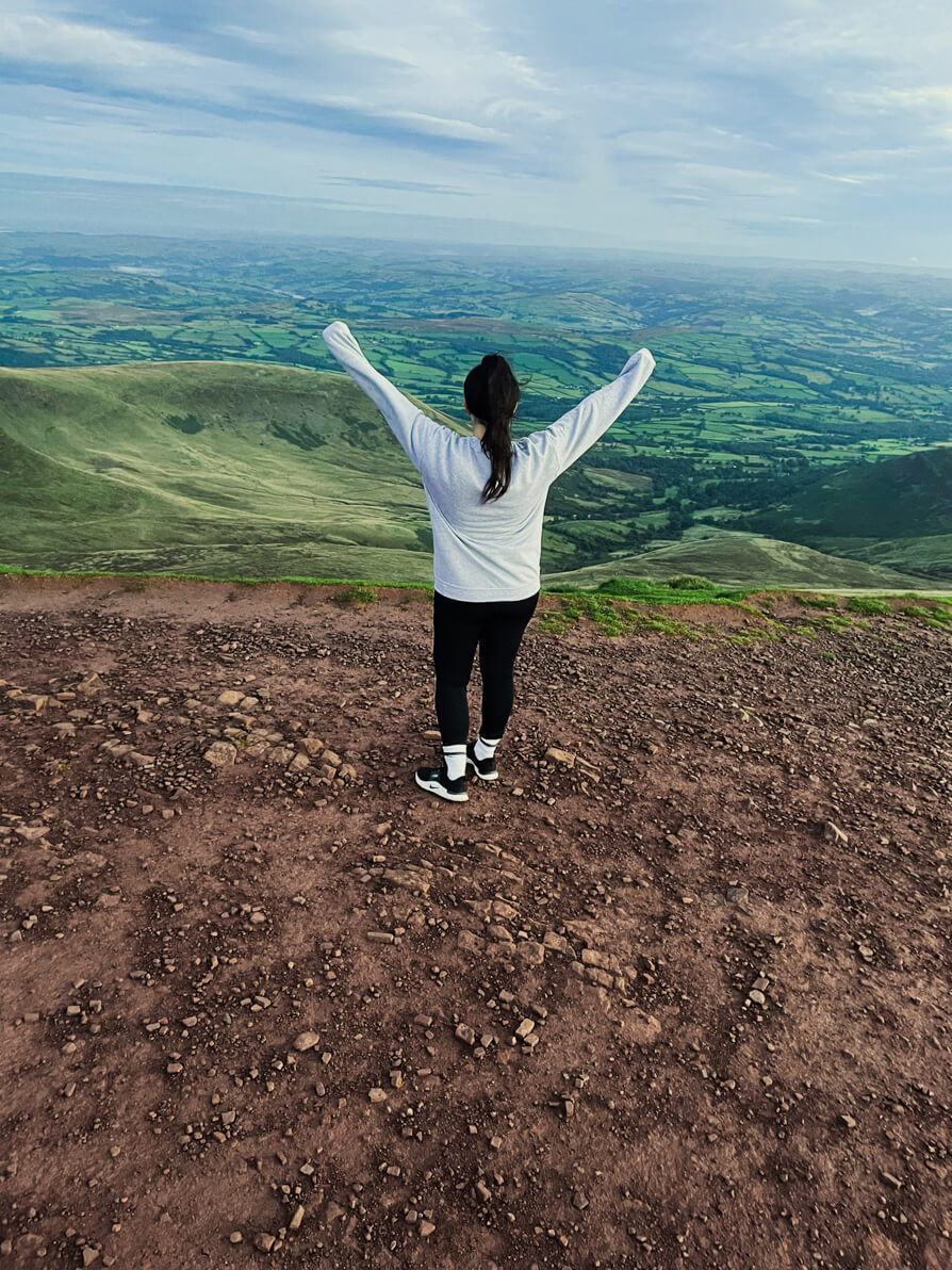 Shireen looking out over the view of Brecon Beacons from Pen Y Fan with arms in the air