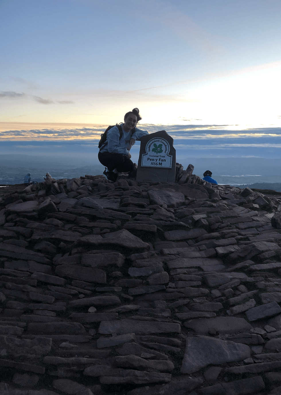 Shireen kneeling next to the Pen Y Fan National Trust sign with sun rising in back