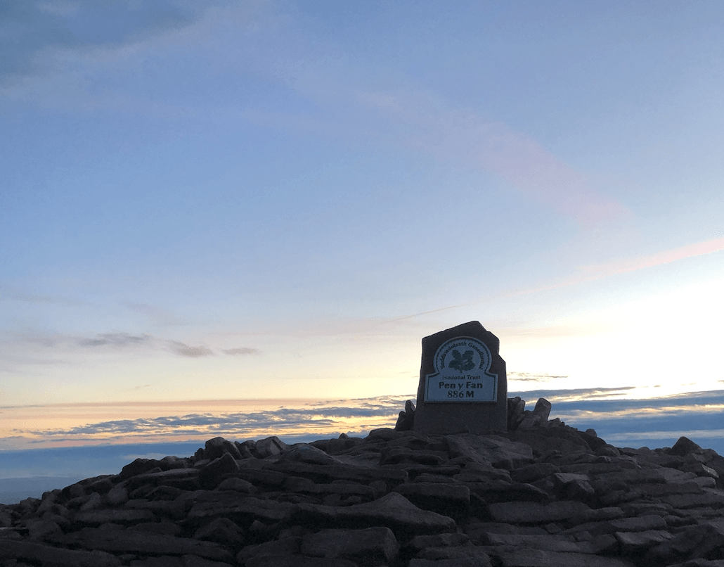 Image of Pen Y Fan National Trust sign and plaque at the top of Pen Y Fan mountain with sun rising in the background