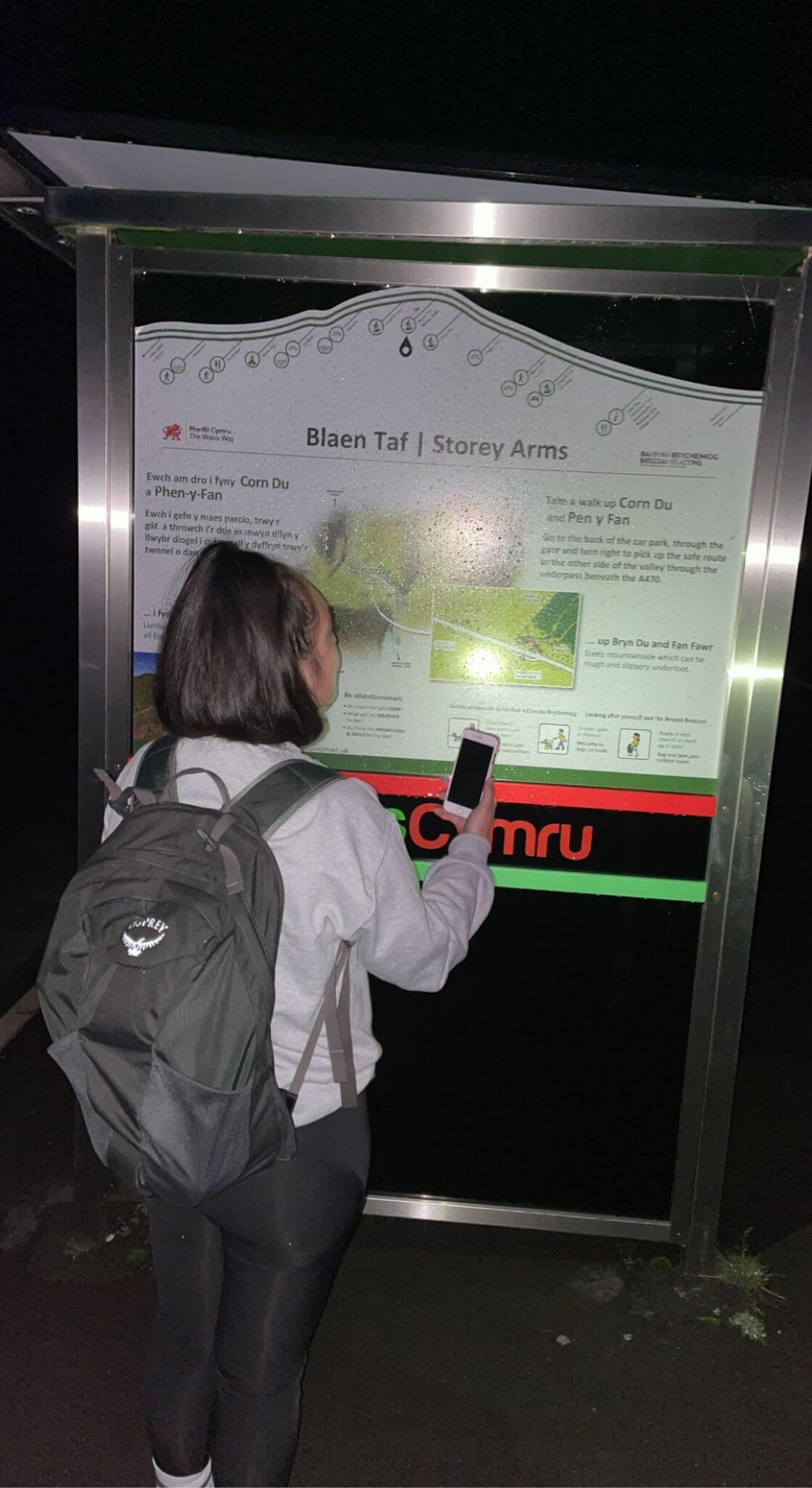 Shireen looking at Storey Arms Centre information board using her phone torch for light. Shireen has an Osprey backpack on