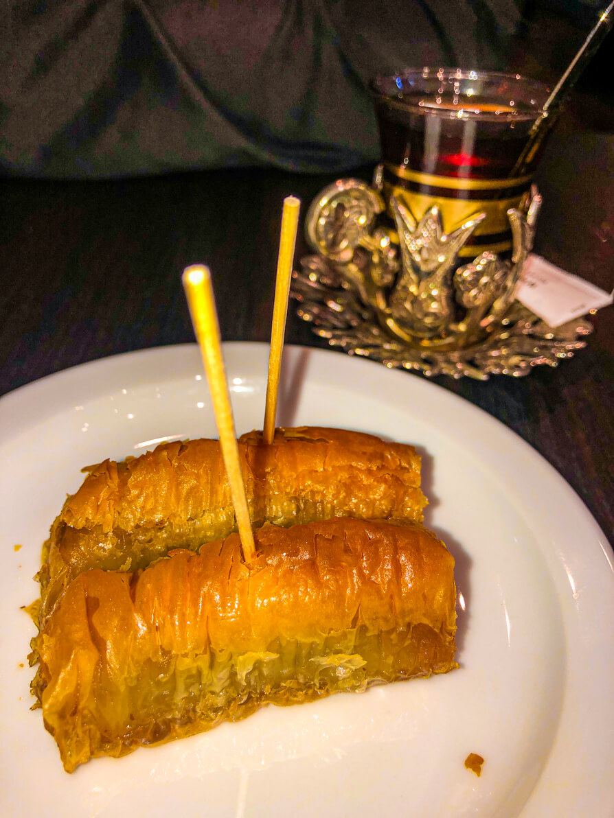 Two small baklava pieces with sticks in the centre on a white plate and Turkish tea cup in the background. Both on a black table in Saray restaurant in Cardiff, Wales