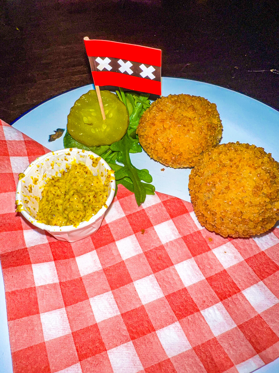 Bitterballen and pickles on a white plate and red and white napkin with Amsterdam flag on a stick in the centre on Amsterdam food tour with Eating Europe