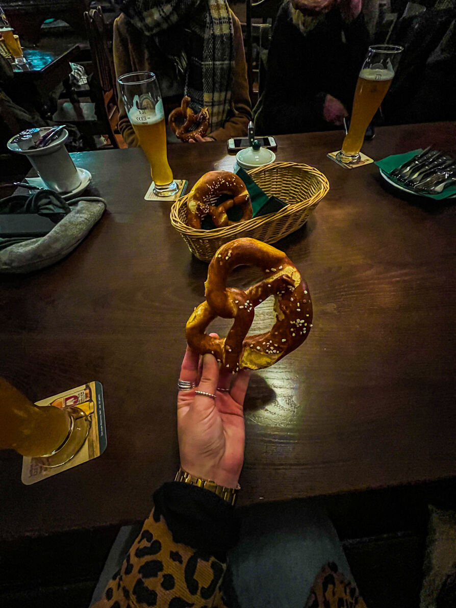 Image of Shireen's left hand holding one pretzel up in from of pretzels on a green napkin in a woven basket on a brown table in Munich Germany