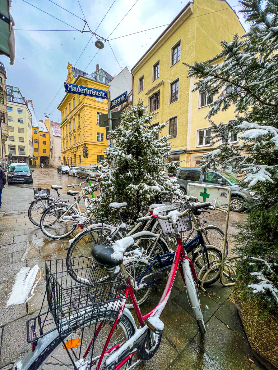 Image of snow-capped Munich street including bikes and trees with yellow buildings in background