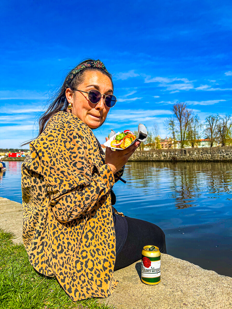 Image of Shireen holding baguette and looking excited to eat sitting on the water's edge of rer Vltava with a can of Staropramen in the forefront
