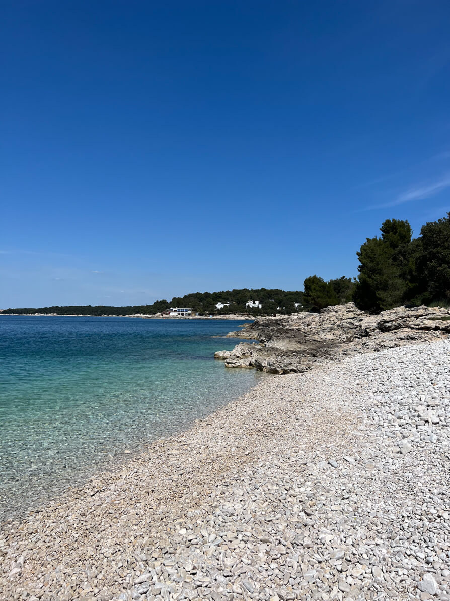 View of the right side of Ambrela Beach in Pula Croatia. Pebbles to the right and blue ocean to the left with woods and rocks in the background