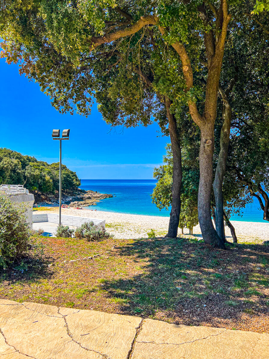 View of Ambrela Beach from the path. Grass, trees and lampost at the front and beach in the background 