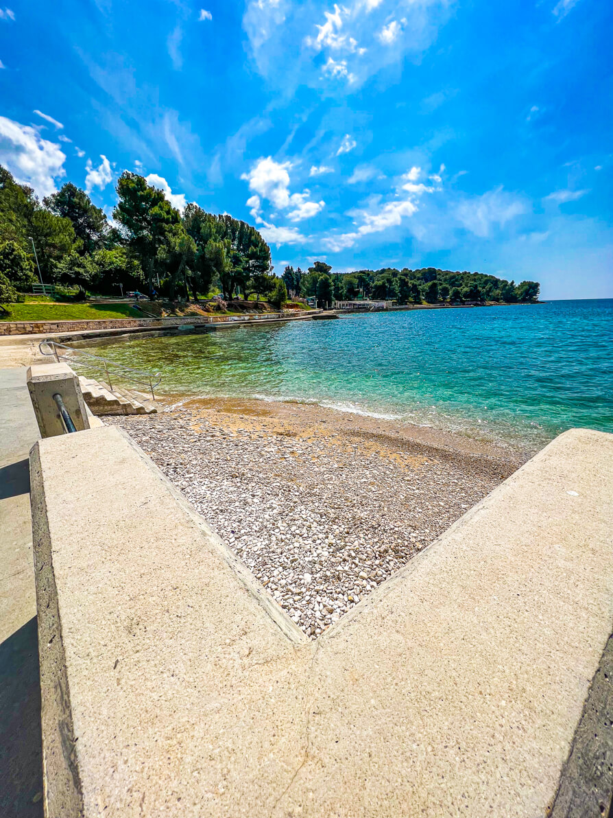 Valkane Beach in Stoja Pula Croatia from the right hand side looking over the wall, pebbles and water. Woodlands in the back.