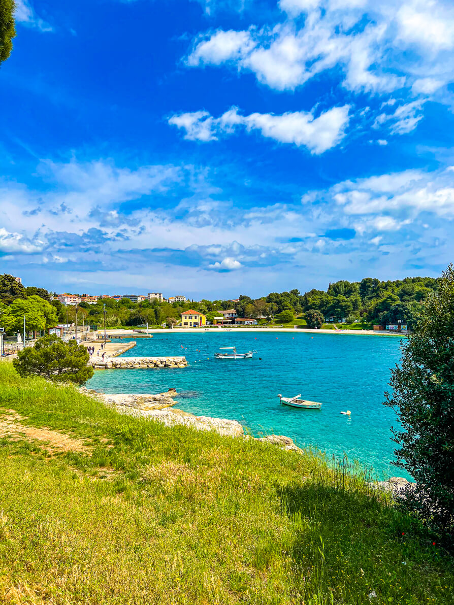 image of Valsaline Beach in Pula Croatia. Grass in the forefront, blue ocean in the back with two boats and Valsaline Beach in background