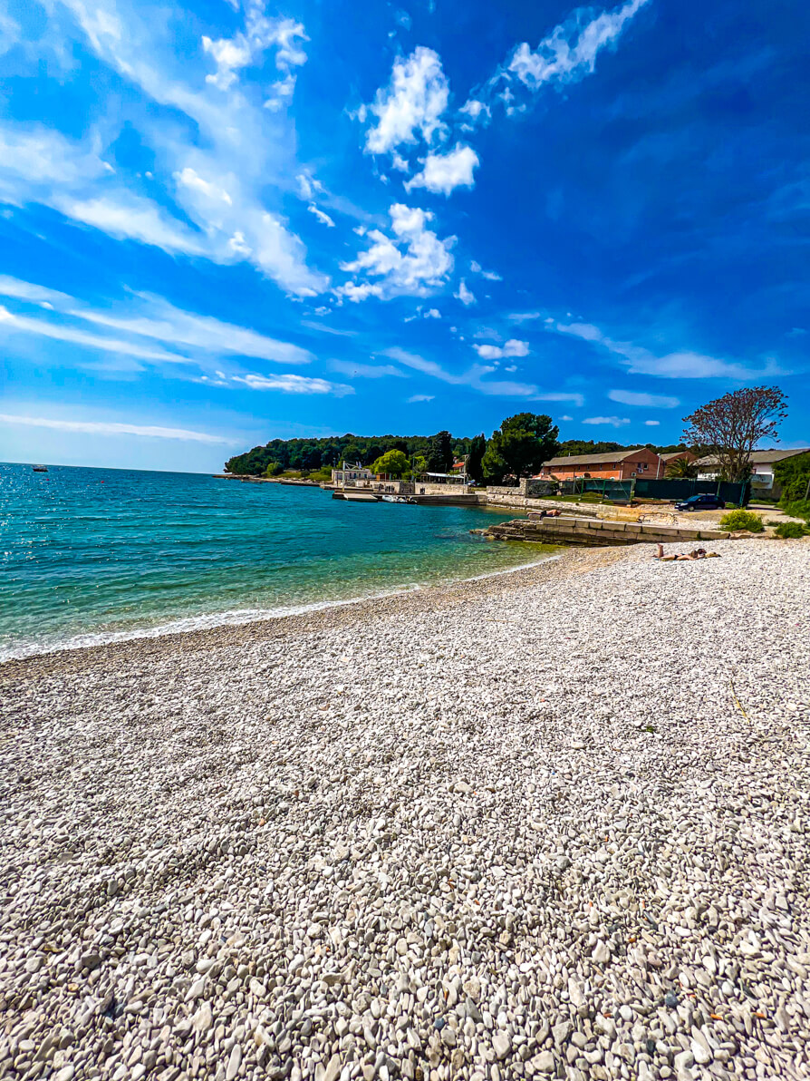 Valsaline Beach in Pula Croatia with pebbles in front, beach, water and woodlands in background from the left hand side of beach