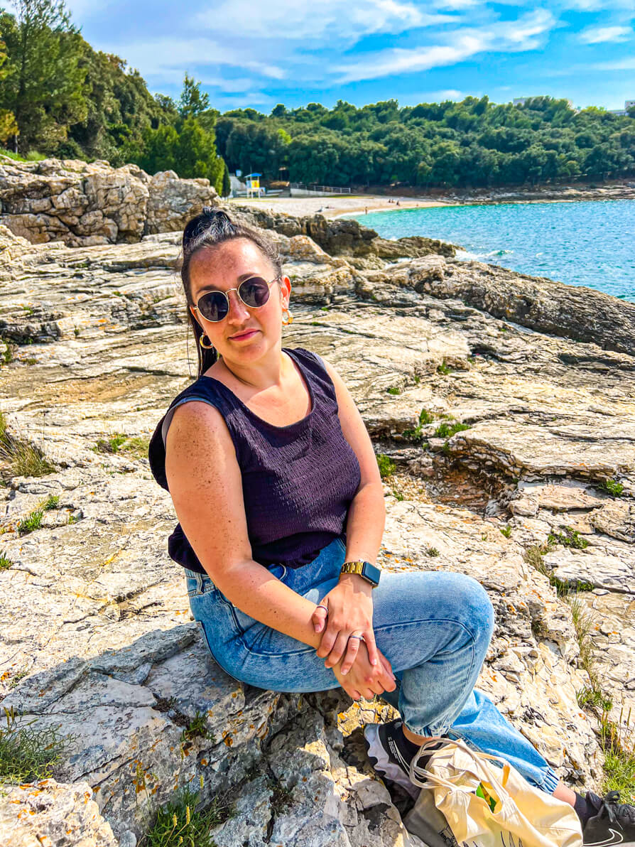 Image of Shireen sitting on the rocks with Pula Beach in background in Stoja Pula Croatia
