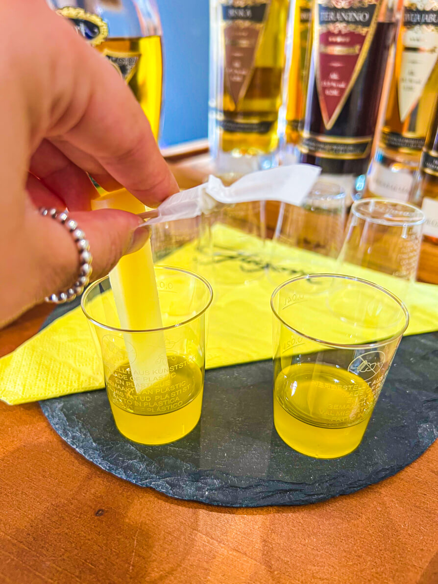 Image of Shireen's left hand dipping Pag cheese into an olive oil sample in a plastic shot cup. Next is another plastic shot cup with a darker olive oil in it and both are on a black slate in Zadar Croatia