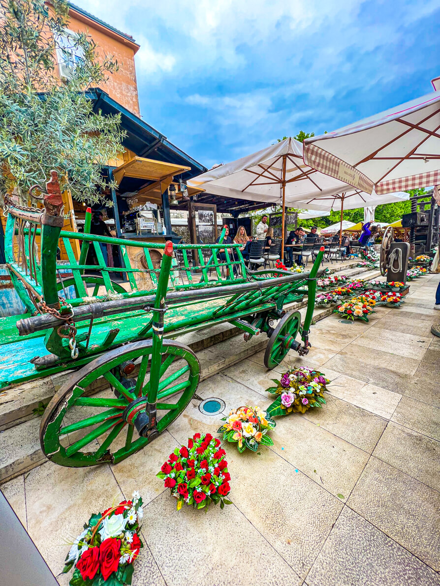 Image of a traditional green cart where fruits and vegetables would be carried in the Green Market in Zadar Croatia. In front are four baskets of flowers and in the back is the market cafe.