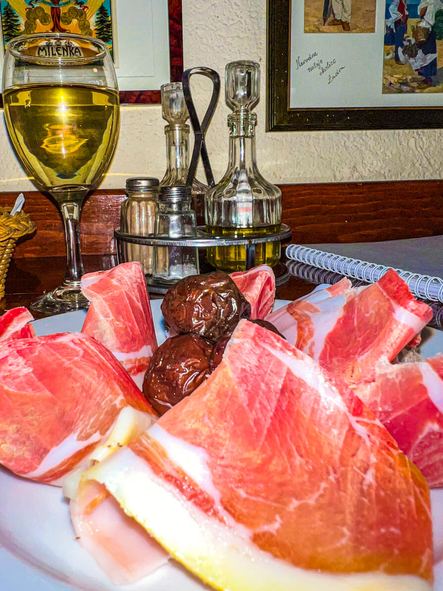 Up close image of Dalmatian Prosciutto with dried, purple olives in the middle on a white place with white wine in the background in Zadar Croatia