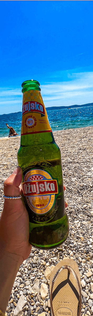 Close up image of Shireen's left hand holding up a glass bottle of Ozujsko at the beach in Croatia