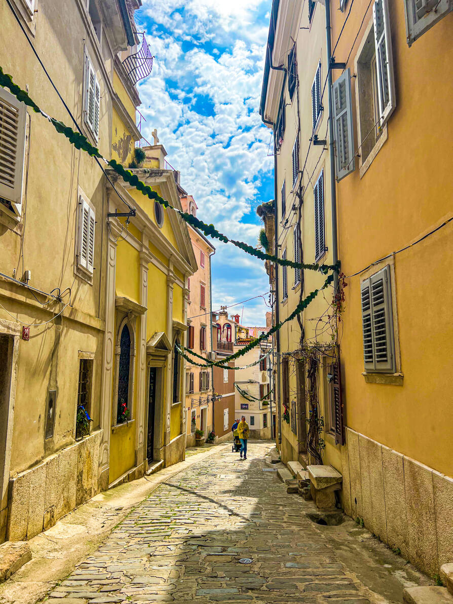 Image of cobbled alleys in Piran town, buildings either side are yellow with bunting across with blue sky in background