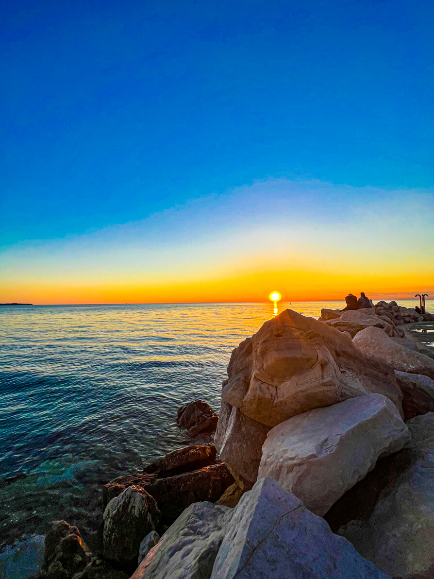 Image of sunset in Piran, rocks and sea in forefront and orange sun going down against blue sky in back