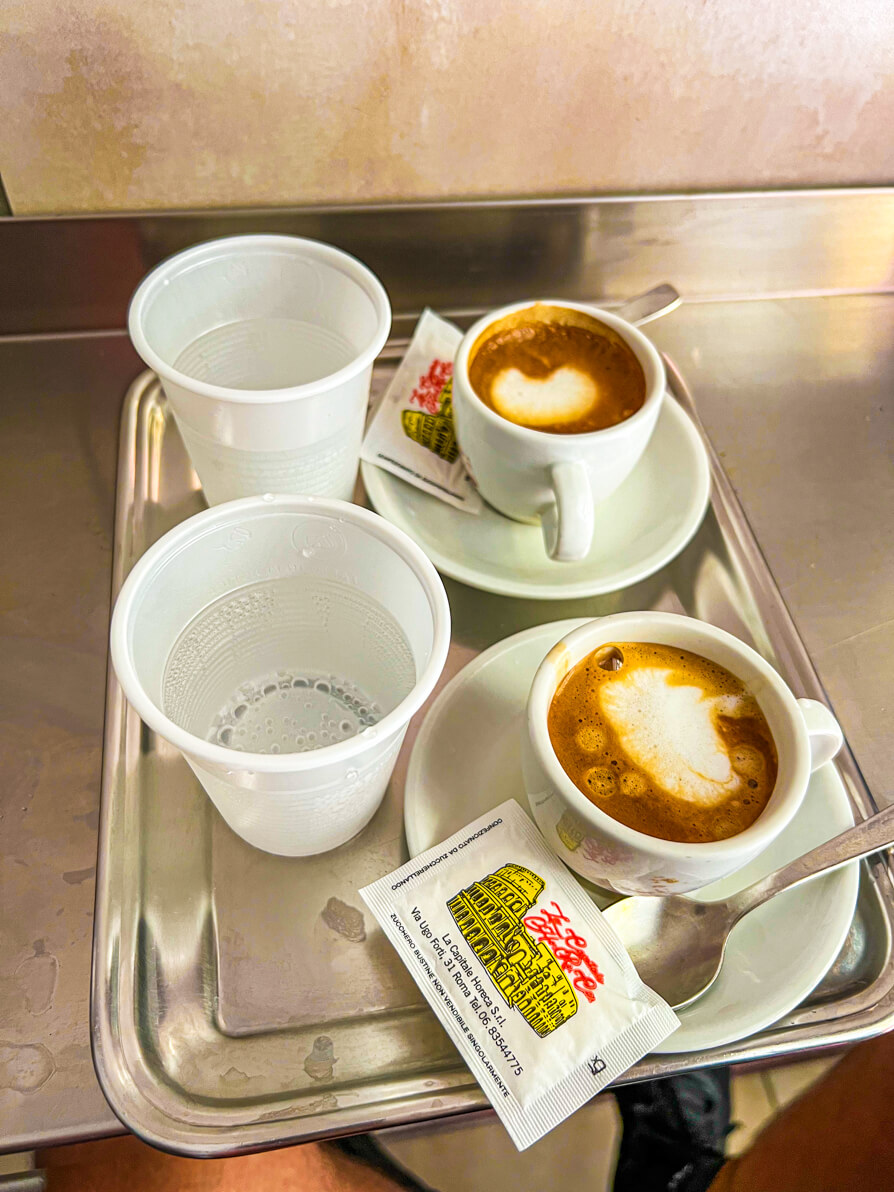 Image of 2 white paper cups with water and 2 caffe lattes in white mugs on a silver tray in Naples Italy