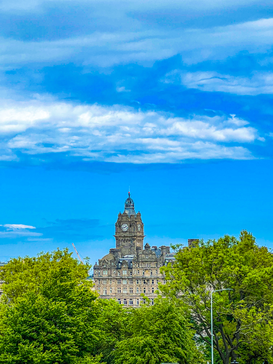 Image of Balmoral Hotel from a distance looking down. The hotel is peering through green trees and the blue sky in back in Edinburgh