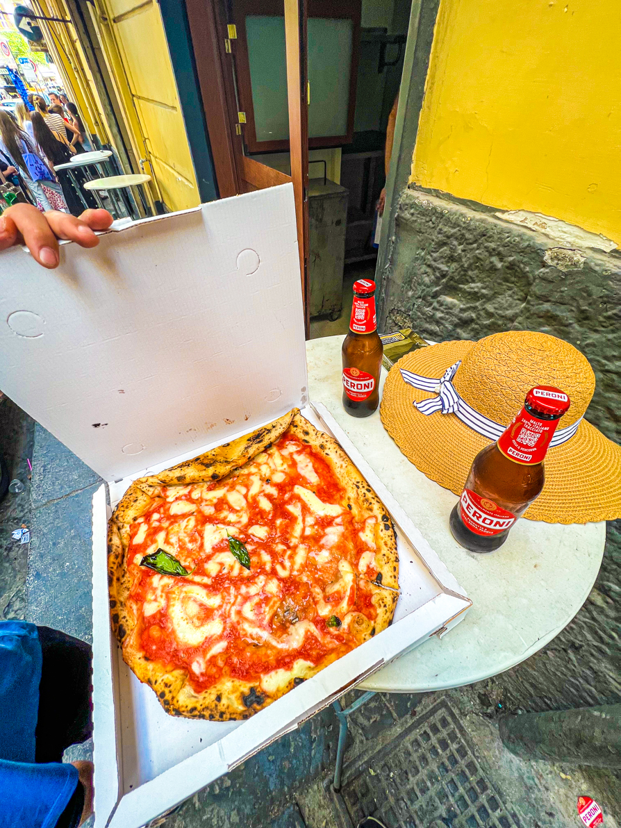 Image of margarita pizza in white box with two bottles of peroni and a straw hat on a white table outside Michele da Pizzeria in Naples Italy