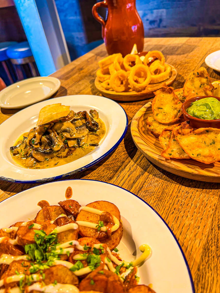 Image of four dishes in Curado Cardiff. Front plate of Patatas Bravas, middle left is Garlic Mushrooms, middle right is prawn tortilla fritters and back plate is Calamari with brown jug in far back.
