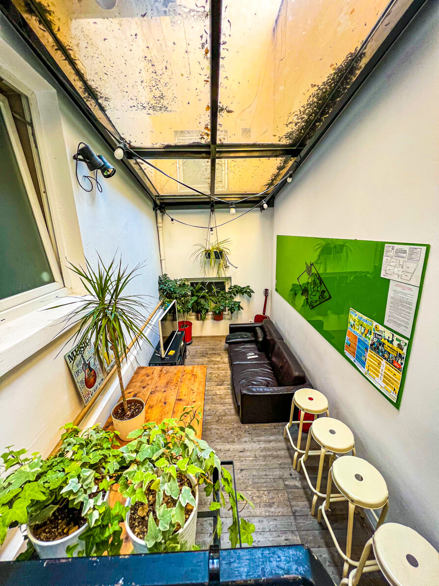 Image of conservatory living area in First Hostel Bucharest. Conservatory has green plants in front with long black settee and tv in back and white stools to the right hand side.