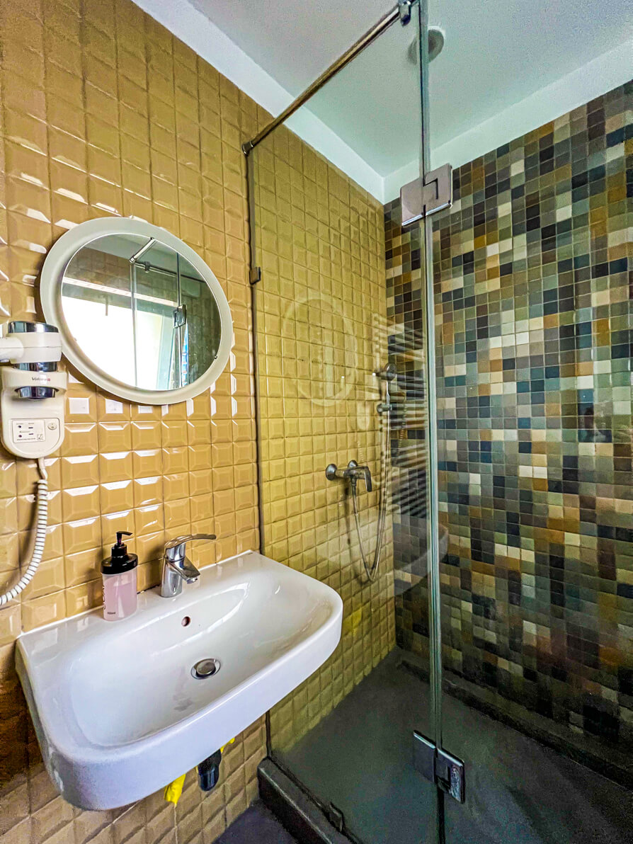 Image of Bathroom in First Hostel in Bucharest, Romania. Bathroom has yellow tiles, circle mirror, white sink and large shower with rectangled tiles