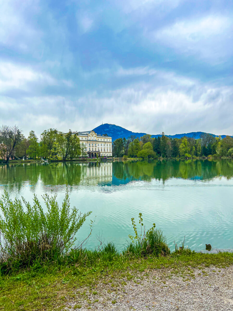 Image of Schloss Leopoldskron Palace, Sound of Music filming location. Image of large white castle in the back left of picture, trees back right and mountains far back with lake in front with reflection of castle. Salzburg, Austria