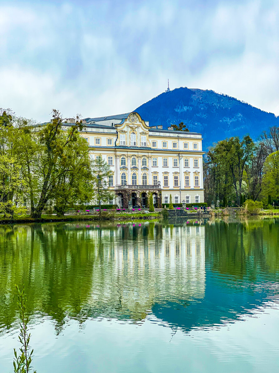 Image of Schloss Leopoldskron Palace, Sound of Music filming location. Image of large white castle in the back left of picture, trees back right and mountains far back with lake in front with reflection of castle. Salzburg, Austria