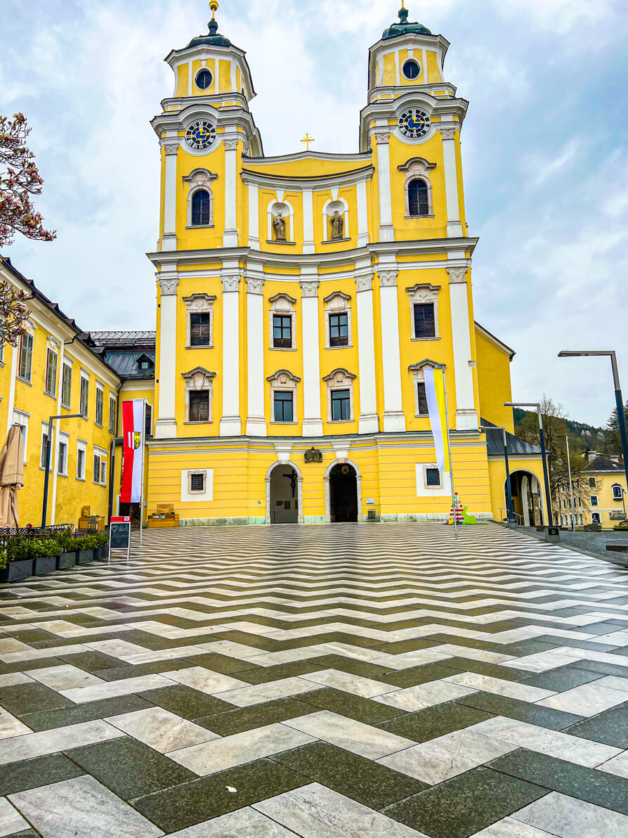 Image of St Michael's Church exterior in Mondsee, Austria with black and white marble floor in front and large, tall, yellow church in background