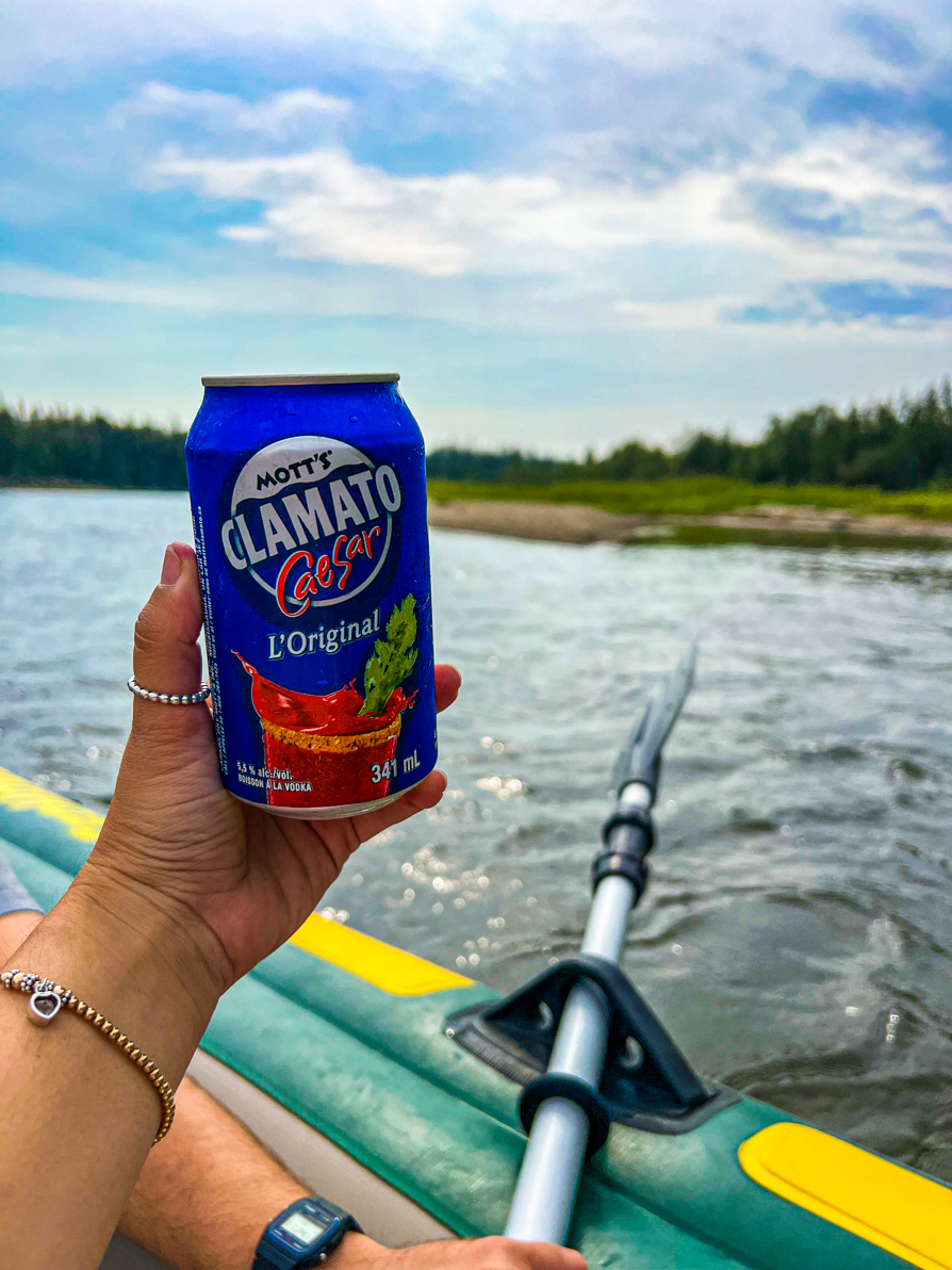 Image of Shireen's left hand holding up a can of Clamato Caesar while on the raft on Red Deer River with river and paddle in background
