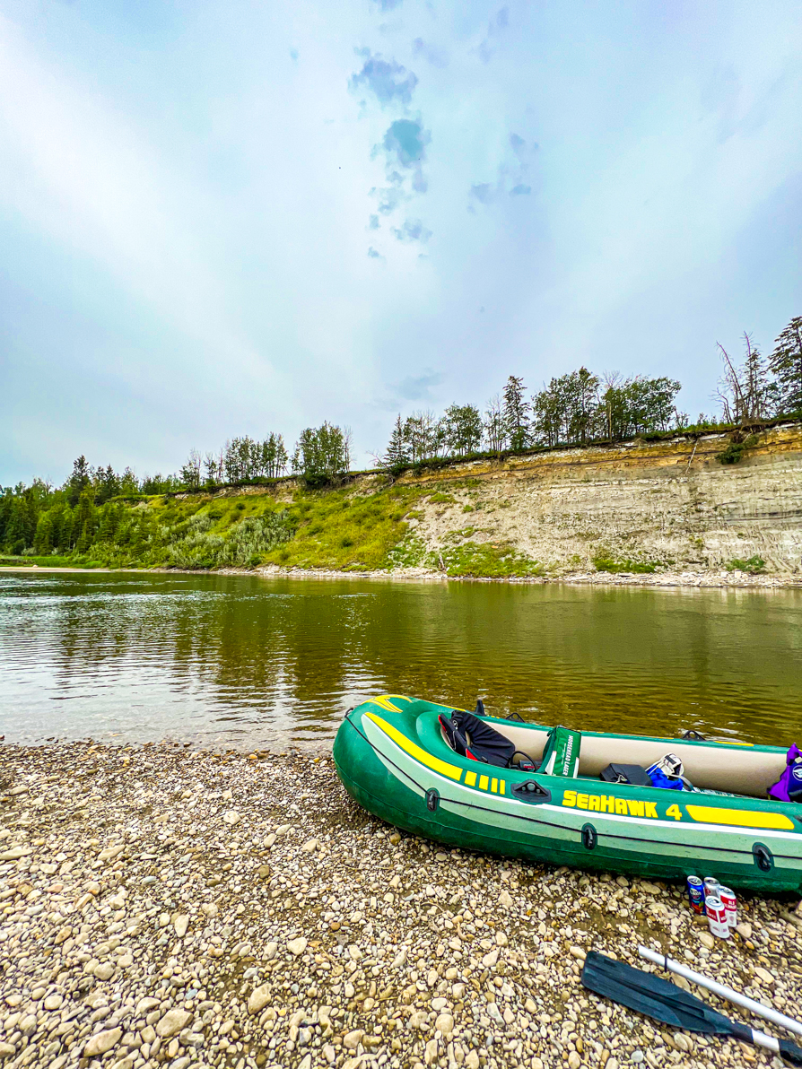 Image of boat and paddle on bank of river with Red Deer river and trees behind in AB