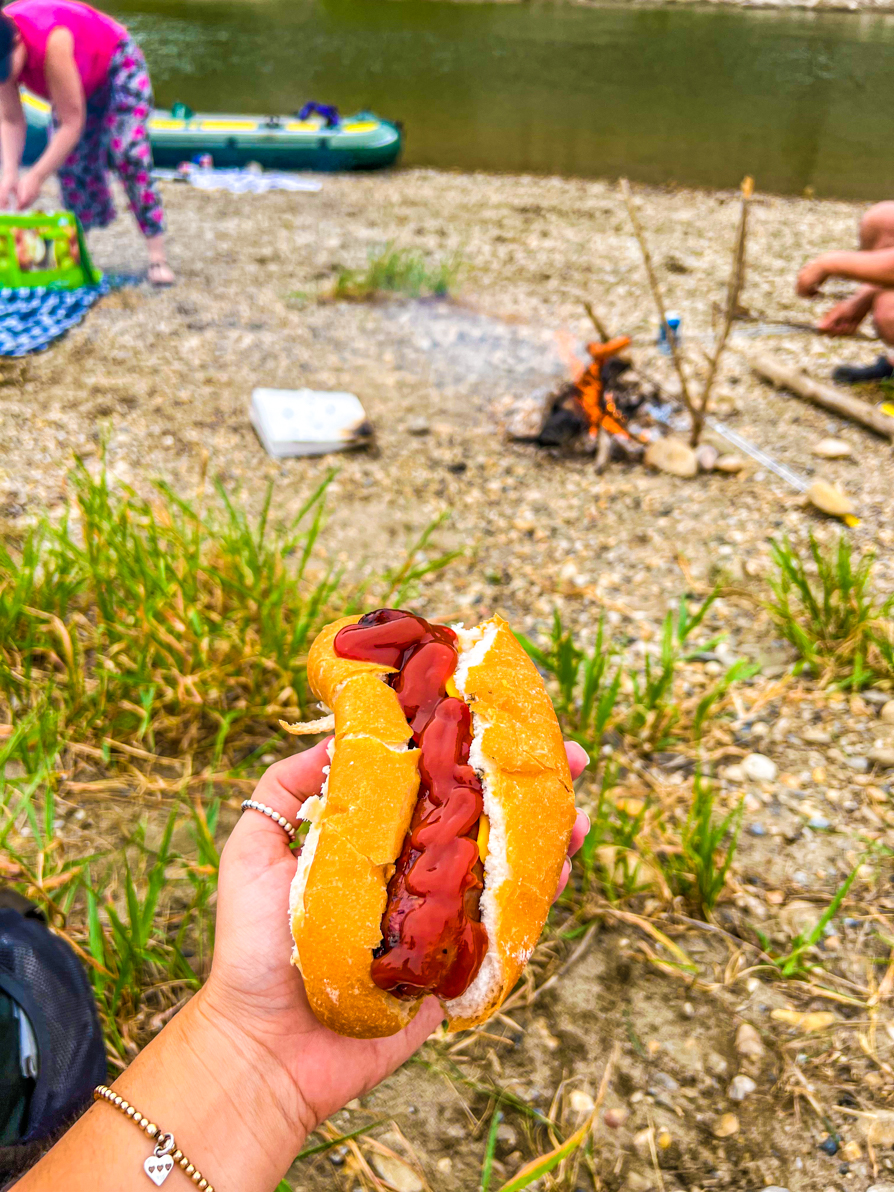 Image of Shireen's left hand holding a hot dog up in front of the fire and river in Red Deer AB Canada