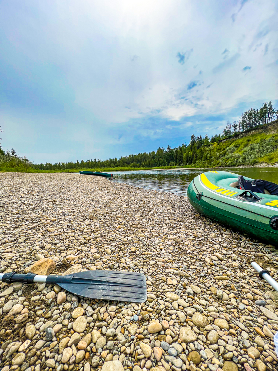 Image of inflatable boat and paddle on the bank of the Red Deer river with river and trees in background. AB Canada