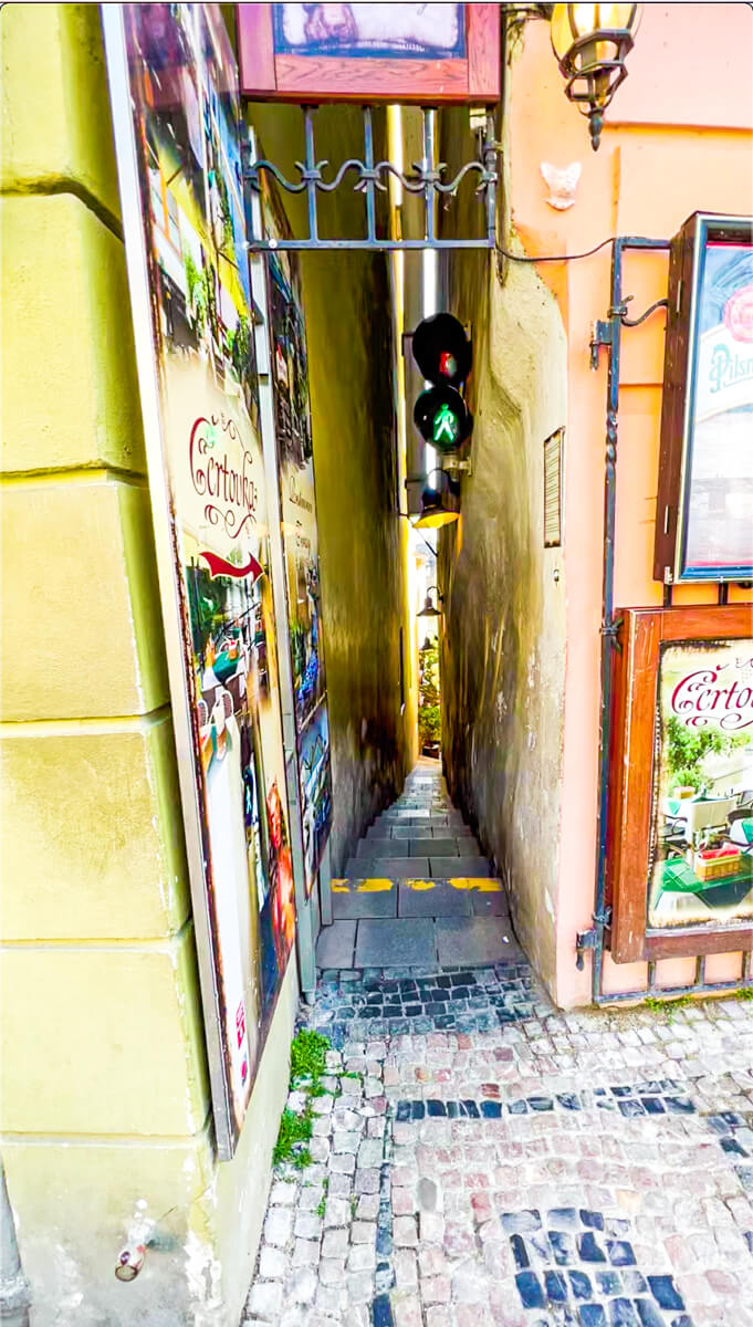 Image of the most narrow street in Prague from the main road view or traffic light showing green and either side buildings are in the shot to show how narrow the street is.