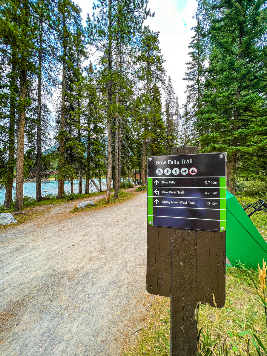 Image of information board about bow falls trail on right of picture with pathway in centre and green trees and blue river to the left in Banff Canada